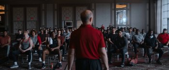Movie still from “National Champions” (2021), directed by Ric Roman Waugh – A group of people sitting in a room with a man standing in front of them; Wide shot, Over the shoulder angle