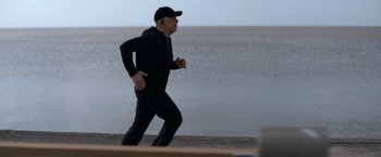 Movie still from “National Champions” (2021), directed by Ric Roman Waugh – A man in a black hat is running by the water; Medium shot, High angle