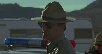 Movie still from “Near Dark” (1987), directed by Kathryn Bigelow – A man in a hat and sunglasses is standing in front of a police car; Close Up shot, Low angle