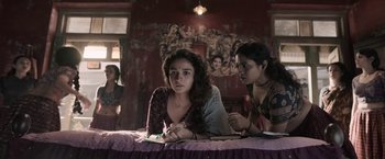 Movie still from “Gangubai Kathiawadi” (2022), directed by Sanjay Leela Bhansali – Two young women sitting at a table in front of a painting; Medium shot, Low angle