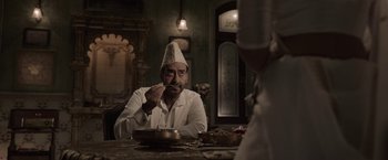 Movie still from “Gangubai Kathiawadi” (2022), directed by Sanjay Leela Bhansali – A man wearing a hat sitting in front of a plate of food on top of a table; Medium shot, Over the shoulder angle