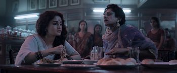 Movie still from “Gangubai Kathiawadi” (2022), directed by Sanjay Leela Bhansali – Two women sitting at a dinner table in front of other women; Medium shot, Over the shoulder angle