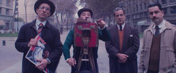 Movie still from “Neruda” (2016), directed by Pablo Larraín – A man is holding something in his mouth while standing next to two other men; Medium shot, Low angle