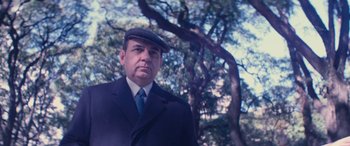 Movie still from “Neruda” (2016), directed by Pablo Larraín – A man wearing a suit and a hat standing in front of a tree; Medium shot, Low angle