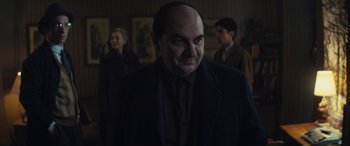 Movie still from “Neruda” (2016), directed by Pablo Larraín – A man in a suit and tie standing in front of a group of people in a room; Medium shot, Over the shoulder angle