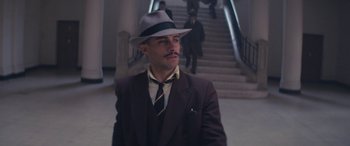 Movie still from “Neruda” (2016), directed by Pablo Larraín – A man in a suit and a hat standing in front of a staircase; Medium shot, Low angle