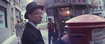 Movie still from “Neruda” (2016), directed by Pablo Larraín – A man in a hat and glasses putting money into a fire hydrant; Medium shot, Low angle