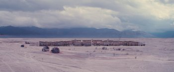 Movie still from “Neruda” (2016), directed by Pablo Larraín – A truck driving down a dirt road next to a bunch of buildings; Extreme Wide shot, High angle