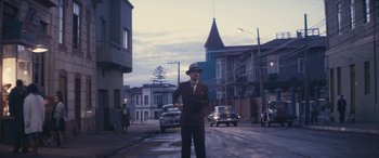 Movie still from “Neruda” (2016), directed by Pablo Larraín – A man standing on the side of the road in front of some cars; Wide shot, Low angle