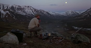 Movie still from “Never Cry Wolf” (1983), directed by Carroll Ballard – A man sitting on a chair in front of an open fire in the mountains; Wide shot, High angle