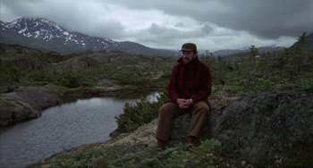 Movie still from “Never Cry Wolf” (1983), directed by Carroll Ballard – A man sitting on top of a rock near a body of water; Wide shot, High angle