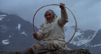 Movie still from “Never Cry Wolf” (1983), directed by Carroll Ballard – An older man holding a hula hoop while sitting on the ground in front of some mountains; Medium shot, Low angle