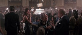 Movie still from “Never Say Never Again” (1983), directed by Irvin Kershner – An old woman smoking a cigarette in a crowded room; Medium shot, Over the shoulder angle