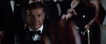Movie still from “Never Say Never Again” (1983), directed by Irvin Kershner – A man in a suit and bow tie in front of a group of people; Close Up shot, Over the shoulder angle