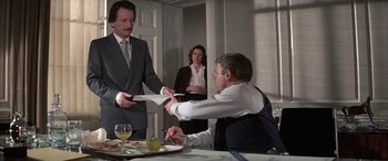 Movie still from “Never Say Never Again” (1983), directed by Irvin Kershner – A man in a business suit is handing a piece of paper to another man in a business suit; Medium shot, Over the shoulder angle