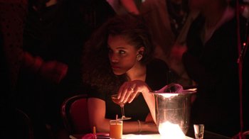 Movie still from “New Jack City” (1991), directed by Mario Van Peebles – A woman sitting at a table with a drink in front of her; Medium shot, Over the shoulder angle