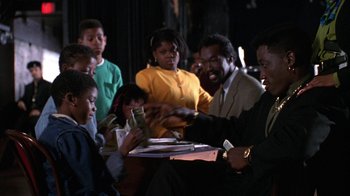 Movie still from “New Jack City” (1991), directed by Mario Van Peebles – A group of people sitting around a table with money; Medium shot, Over the shoulder angle