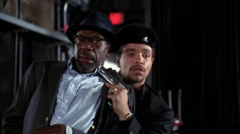 Movie still from “New Jack City” (1991), directed by Mario Van Peebles – A man holding a gun next to another man in a hat; Medium shot, Low angle