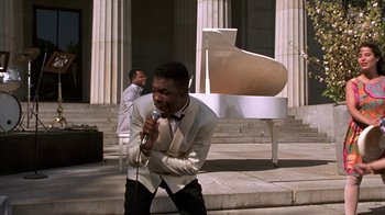 Movie still from “New Jack City” (1991), directed by Mario Van Peebles – A man in a white jacket singing into a microphone; Medium shot, High angle