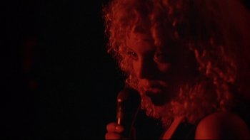 Movie still from “New Rose Hotel” (1998), directed by Abel Ferrara – A man with curly red hair holding a microphone in a dark room; Close Up shot, Low angle