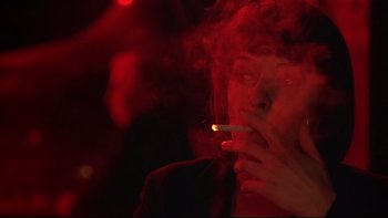 Movie still from “New Rose Hotel” (1998), directed by Abel Ferrara – A woman smoking a cigarette in a dark room; Close Up shot, Over the shoulder angle