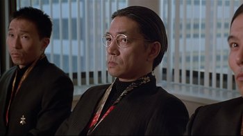 Movie still from “New Rose Hotel” (1998), directed by Abel Ferrara – A man in a black suit and glasses looks to his left; Close Up shot, Over the shoulder angle