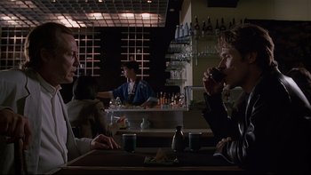 Movie still from “New Rose Hotel” (1998), directed by Abel Ferrara – A group of people sitting at a table drinking; Medium shot, Over the shoulder angle
