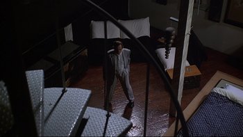 Movie still from “New Rose Hotel” (1998), directed by Abel Ferrara – A man standing on a wooden floor in front of a couch; Wide shot, Overhead angle