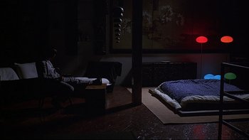 Movie still from “New Rose Hotel” (1998), directed by Abel Ferrara – A man sitting in a chair in front of a bed in a dark room; Wide shot, High angle