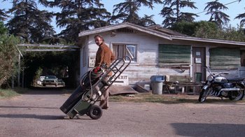 Movie still from “Night Moves” (1975), directed by Arthur Penn – A man is carrying a dolly with a guitar case on it; Wide shot, Low angle