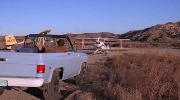 Movie still from “Night Moves” (1975), directed by Arthur Penn – A man driving a truck in front of an airplane on a dirt road; Wide shot, Low angle