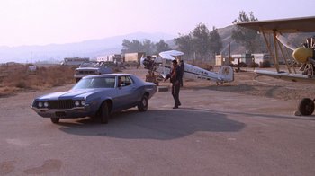 Movie still from “Night Moves” (1975), directed by Arthur Penn – A man standing next to a car and a plane; Wide shot, High angle