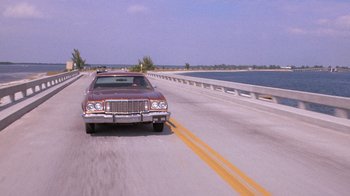 Movie still from “Night Moves” (1975), directed by Arthur Penn – An old car driving down a road next to a body of water; Extreme Wide shot, Over the shoulder angle