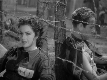Movie still from “Night Train to Munich” (1940), directed by Carol Reed – A woman and a man sitting next to each other in front of a barbed wire fence; Medium shot, Low angle
