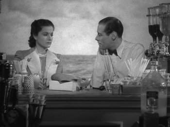 Movie still from “Night Train to Munich” (1940), directed by Carol Reed – A man and a woman sitting at a table in front of the ocean; Medium shot, High angle