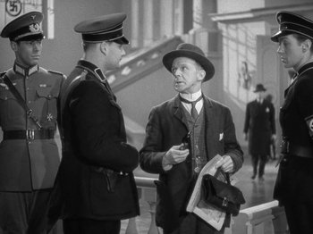 Movie still from “Night Train to Munich” (1940), directed by Carol Reed – A man in a suit and hat talking to another man in a suit and hat; Medium shot, Over the shoulder angle