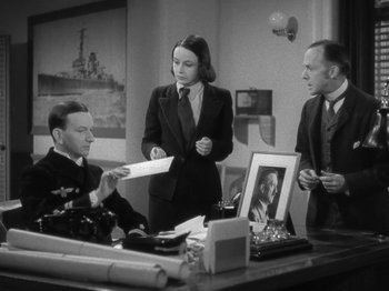 Movie still from “Night Train to Munich” (1940), directed by Carol Reed – A man and two women are standing in front of a desk; Medium shot, High angle