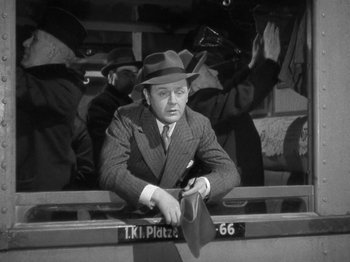 Movie still from “Night Train to Munich” (1940), directed by Carol Reed – A man in a suit and a hat sitting on a bench; Medium shot, Low angle