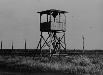 Movie still from “Night and Fog” (1956), directed by Alain Resnais – A black and white photo of a guard tower in the middle of a field; Extreme Wide shot, Low angle