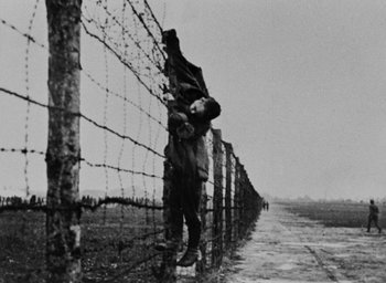 Movie still from “Night and Fog” (1956), directed by Alain Resnais – A man standing next to a barbed wire fence; Wide shot, Low angle