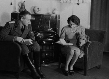 Movie still from “Night and Fog” (1956), directed by Alain Resnais – A man and a woman sitting in a living room with a dog; Medium shot, High angle