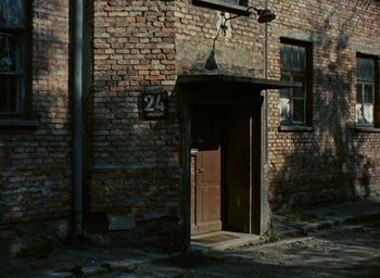 Movie still from “Night and Fog” (1956), directed by Alain Resnais – An old brick building with an open door; Extreme Wide shot, High angle