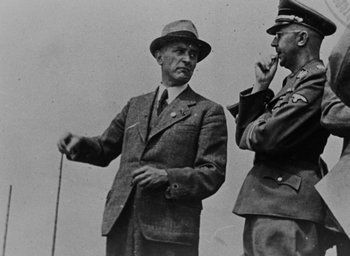 Movie still from “Night and Fog” (1956), directed by Alain Resnais – A man in a suit standing next to another man in a hat; Medium shot, Low angle