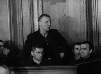 Movie still from “Night and Fog” (1956), directed by Alain Resnais – A black and white photo of a man sitting in a courtroom; Medium shot, Low angle