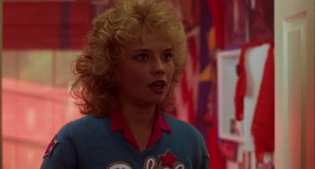 Movie still from “Night of the Comet” (1984), directed by Thom Eberhardt – A woman with blonde curly hair standing in front of a wall; Close Up shot, Over the shoulder angle