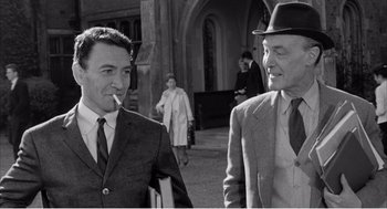Movie still from “Night of the Eagle” (1962), directed by Sidney Hayers – A man in a suit smoking a cigarette next to another man in a suit and hat; Medium shot, High angle
