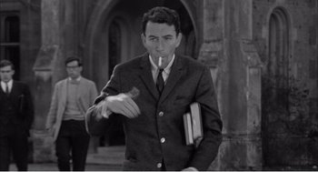 Movie still from “Night of the Eagle” (1962), directed by Sidney Hayers – A black and white photo of a man smoking a cigarette and holding a book; Medium shot, High angle