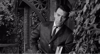 Movie still from “Night of the Eagle” (1962), directed by Sidney Hayers – Black and white photograph of a man in a suit holding a book; Medium shot, Low angle