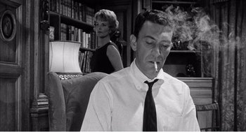 Movie still from “Night of the Eagle” (1962), directed by Sidney Hayers – A man smoking a cigarette in front of a woman in a library; Medium shot, Low angle