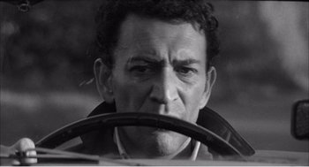 Movie still from “Night of the Eagle” (1962), directed by Sidney Hayers – A man with a beard and a jacket is holding a steering wheel; Close Up shot, Low angle